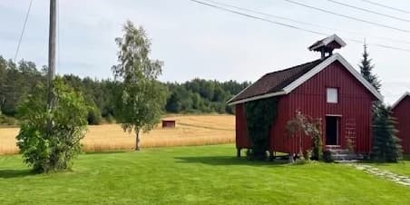 Rødt stabbur på Haughem Gård, med plen foran og kornåker og skog bak
