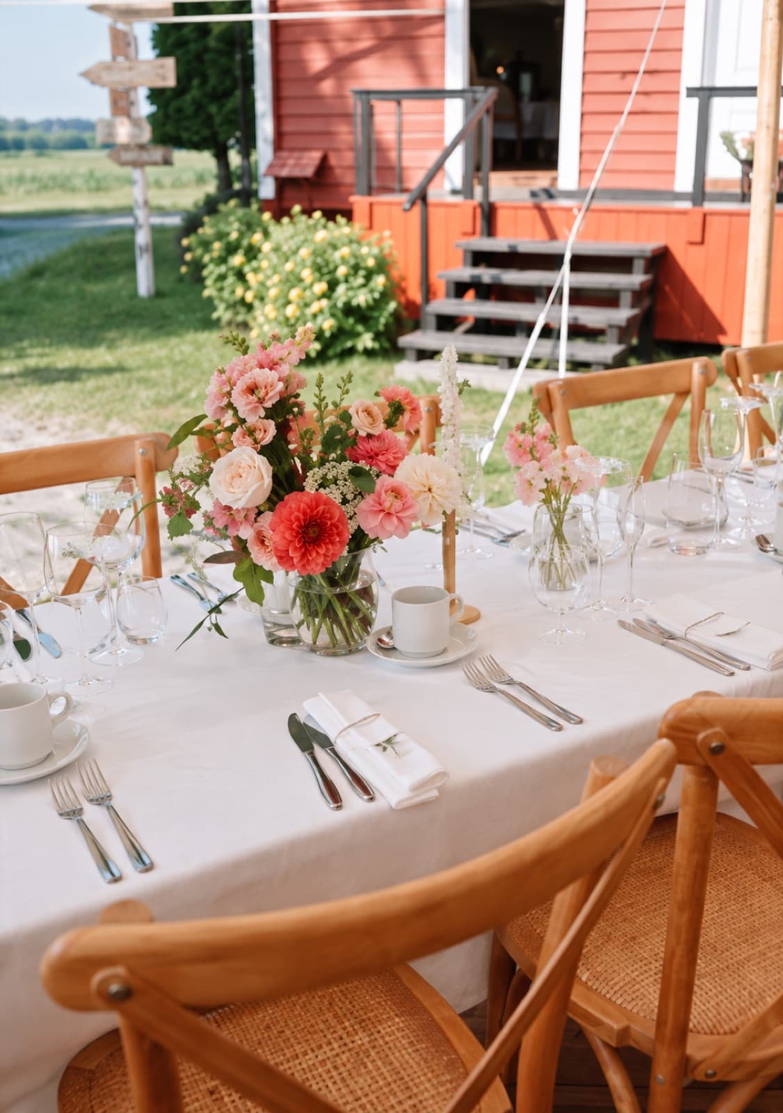 Nærbilde av borddekking med blomsterbukett, bestikk og glass foran det røde gårdshuset