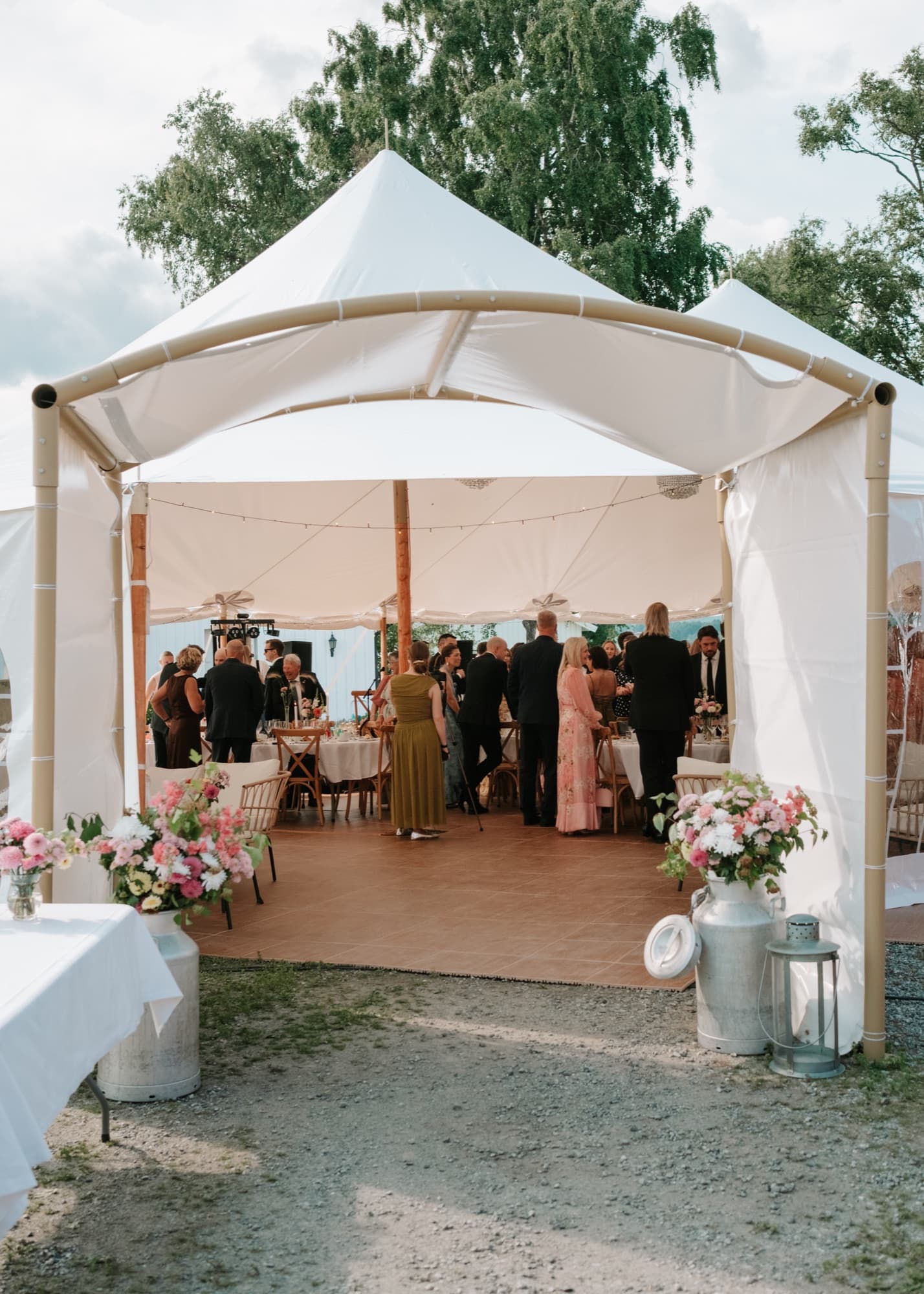 Bryllupsgjester mingler i inngangen til seilduksteltet, med blomster og melkespann på hver side